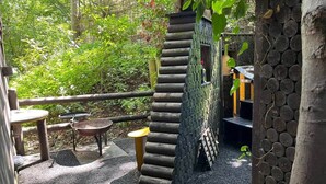 Uma área de estar ao ar livre rústica, com uma roda de água de pedra, mesas de madeira e um cenário natural.