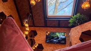 Quarto aconchegante com piso de madeira, uma TV exibindo uma paisagem natural, uma janela com vista para árvores e uma guirlanda de luzes.