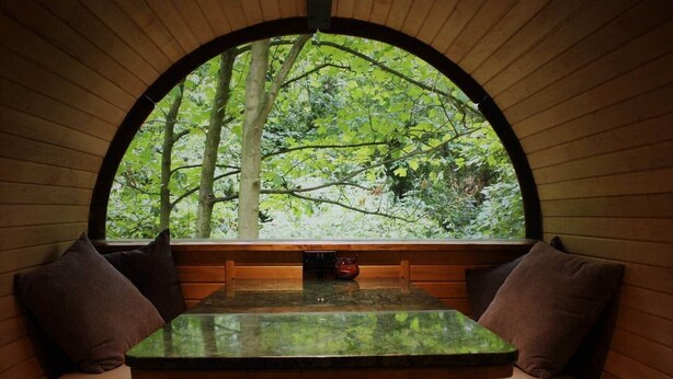 A cozy seating area with a table and benches, a window with a view of a forest, and a small decorative item on the table. - Sleep in the Trees - Hot Tub, Sauna and Slide (Chorleywood)
