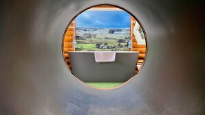 A circular window with a view of a green landscape and a building. - Shepard hut - Hot Tub, Outdoor Shower, Bath (Chorleywood)