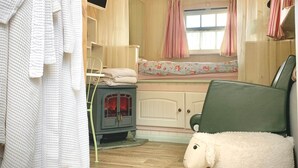 A cozy room with a fireplace, a bed with floral bedding, a green chair, a fluffy white sheep, and a white robe hanging on the wall. - Shepard hut - Hot Tub, Outdoor Shower, Bath (Chorleywood)