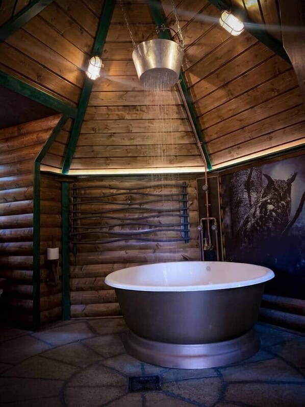 Interior de una cabaña de troncos con una bañera grande, paredes de madera y piso de piedra.