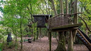A treehouse structure with a wooden staircase and a platform, surrounded by a forested area. - Tree House - Hot Tub on balcony (Chorleywood)