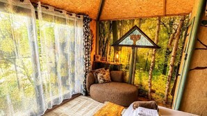 A cozy room with a bed, a wooden bench, and a view of trees through a window. - Tree House - Hot Tub on balcony (Chorleywood)