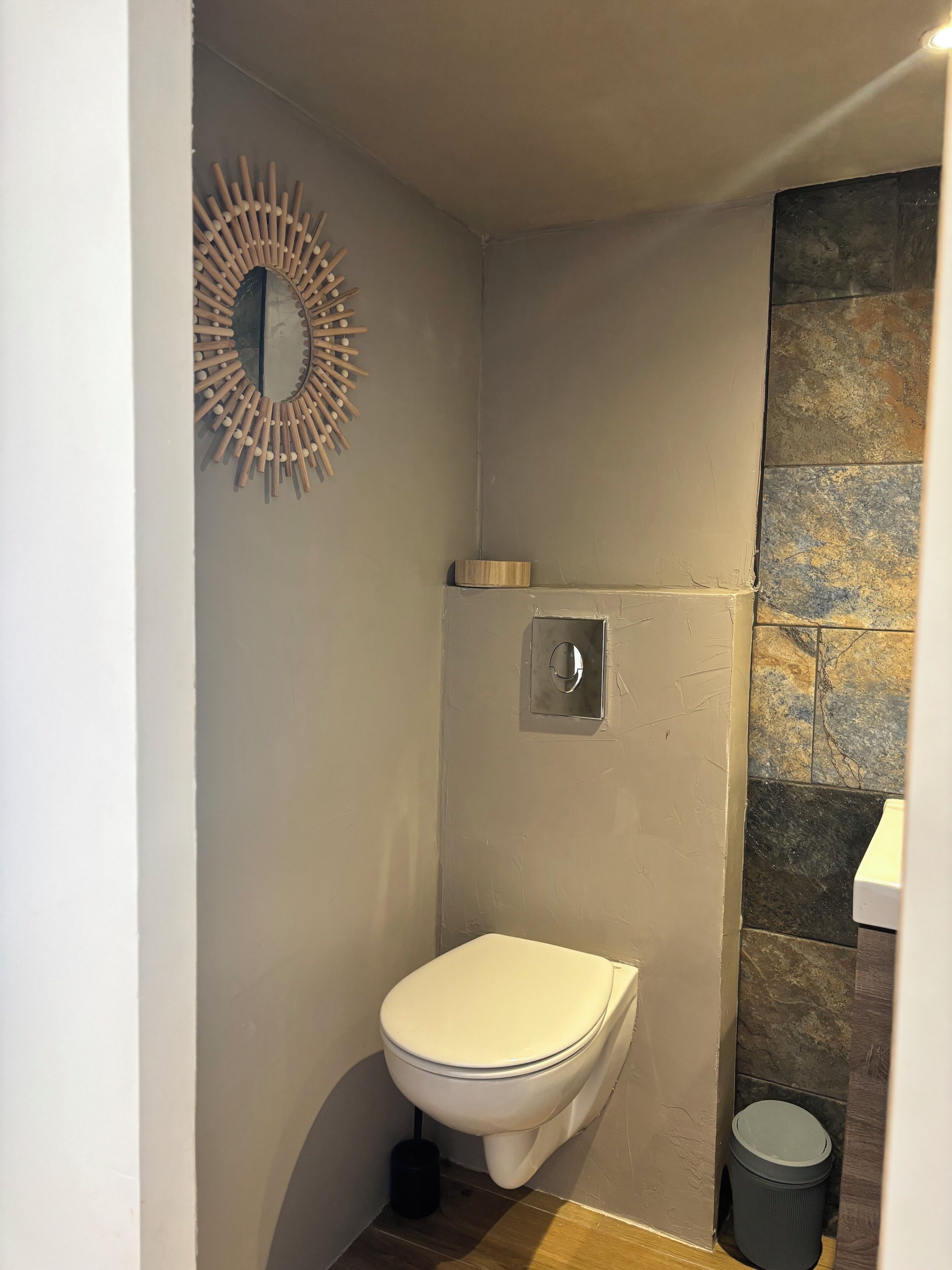 Une petite salle de bain aux dimensions réduites, avec des toilettes blanches, un miroir fixé au mur et une poubelle.