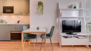 Een moderne keuken met een inbouwoven en magnetron, een eettafel met twee stoelen, een tv op een witte standaard en een plank met decoratieve objecten.