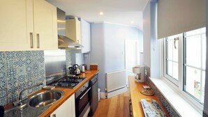 Une cuisine de taille réduite avec des meubles blancs, un évier en acier inoxydable, une cuisinière à gaz et une fenêtre équipée de stores.