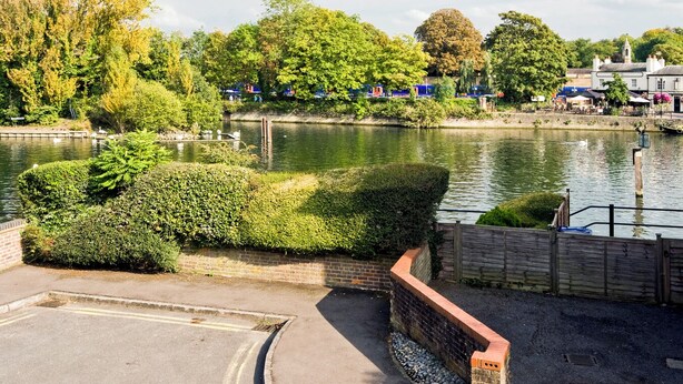 Une promenade au bord de la rivière, bordée d’un mur de briques et de verdure, avec un cours d’eau paisible et des bâtiments en arrière-plan.