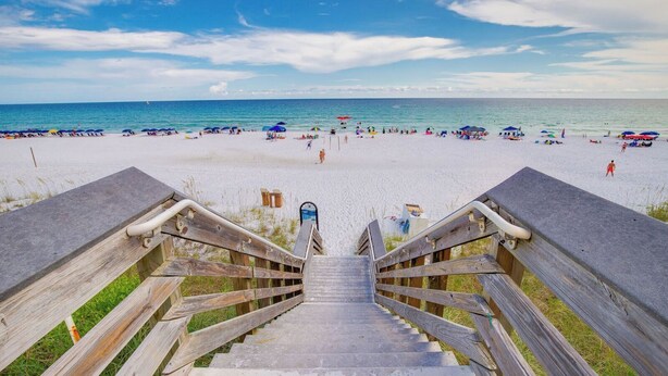 Eine Holztreppe führt zu einem Strand mit Sonnenschirmen und Menschen.