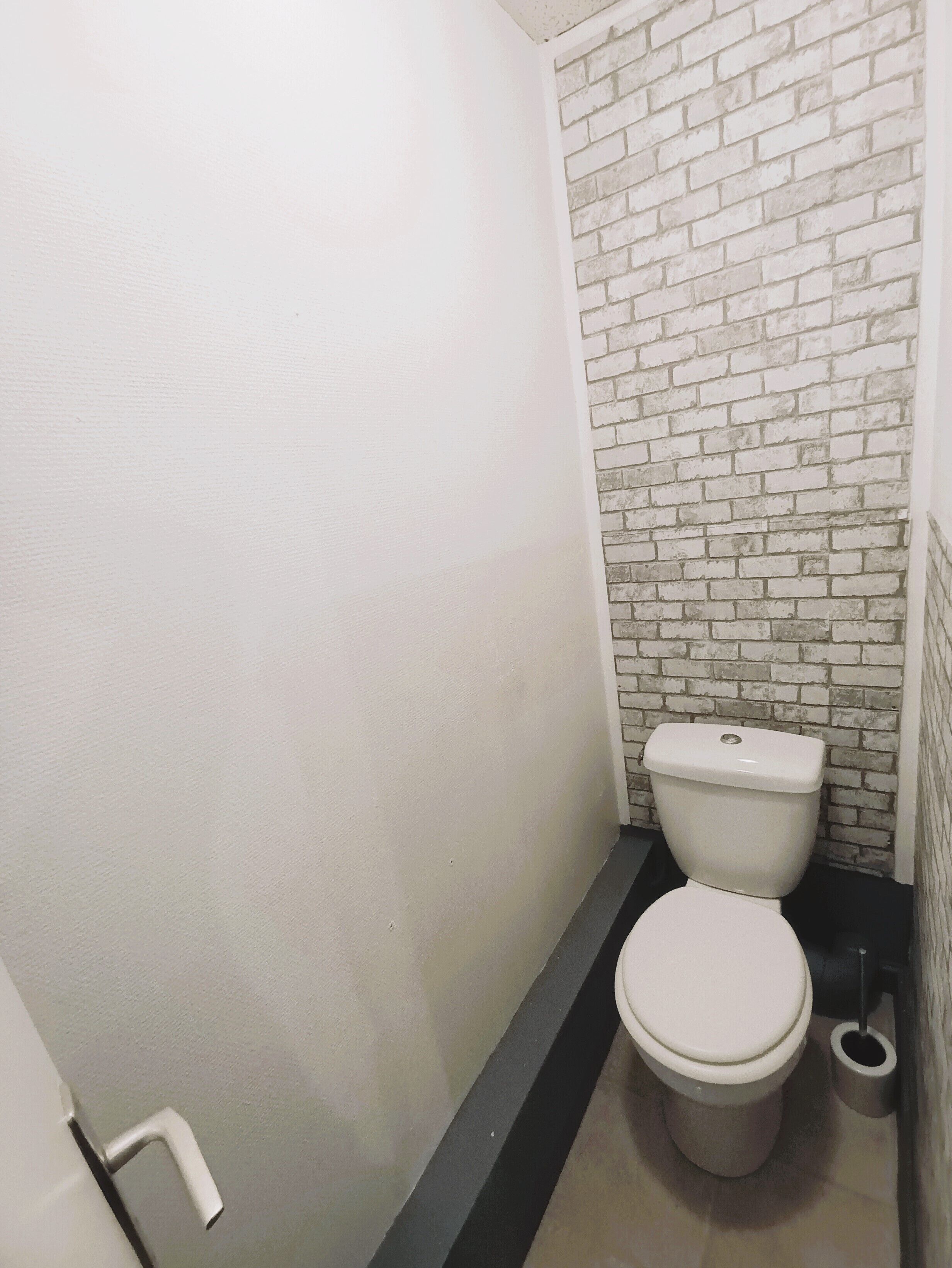 A small, narrow bathroom with a white toilet, a black baseboard, and a white brick wall.