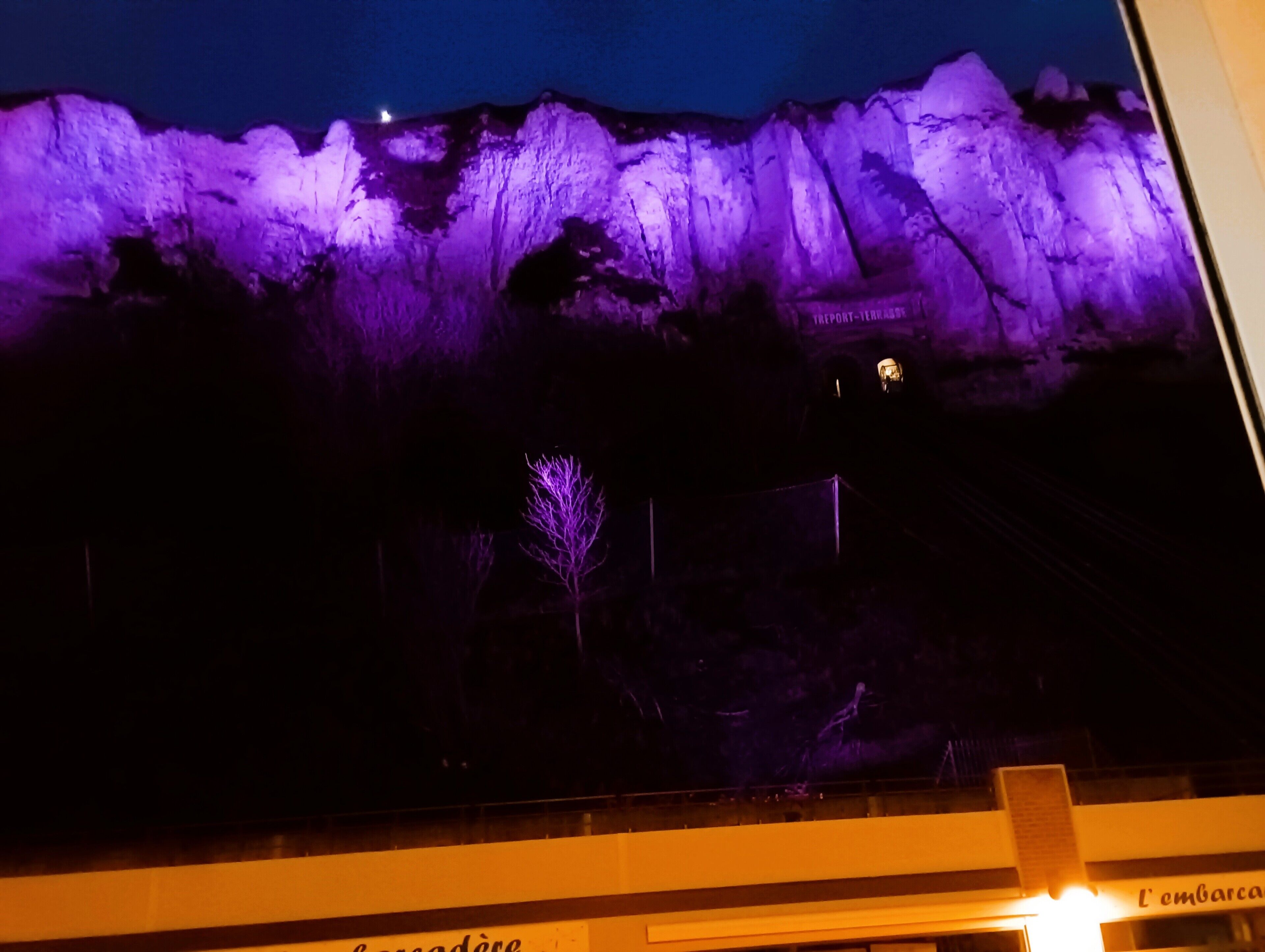 Une vue nocturne d’une montagne illuminée de lumière violette, avec une enseigne indiquant « TRIPORT-TERRASSE », et un arbre au premier plan.