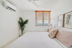 Un dormitorio con cama, ventilador de techo, ventana con persianas, planta en maceta y un cuadro en la pared.