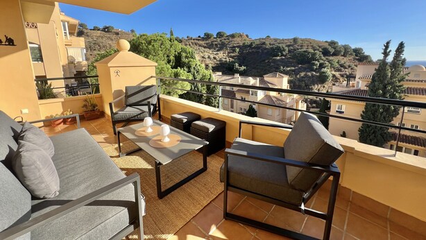 Ein Balkon mit einer Couch, Stühlen und einem Tisch, mit Blick auf eine Hügelkuppe mit Gebäuden und Bäumen.