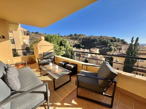 Un balcón con sofá, sillas y mesa, con vistas a una colina con edificios y árboles.