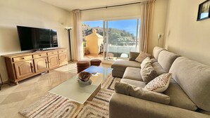Ein Wohnzimmer mit Flachbildfernseher, Holzschrank, beiger Couch und Blick auf einen Balkon mit Geländer und Topfpflanzen.