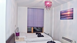 Quarto com duas camas, janela com persianas, aquecedor fixado na parede, abajur e uma pintura de pôr do sol.