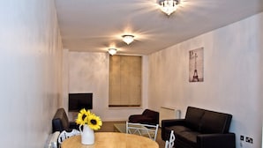 Uma sala de estar com uma mesa de madeira redonda, cadeiras brancas, um sofá preto, uma televisão e uma janela com persianas.