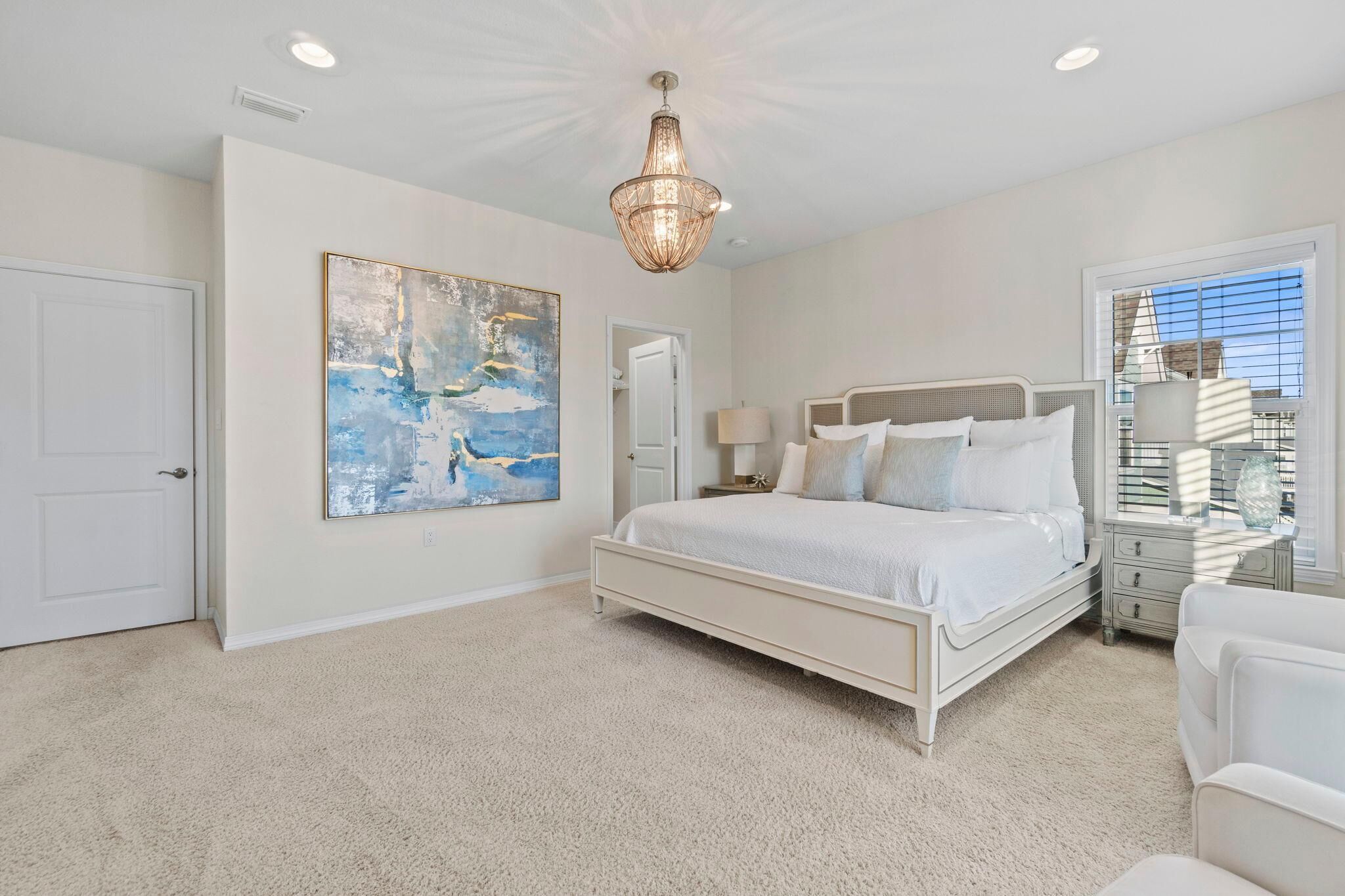 A bedroom with a large bed, a chandelier, a painting, and a window with blinds.