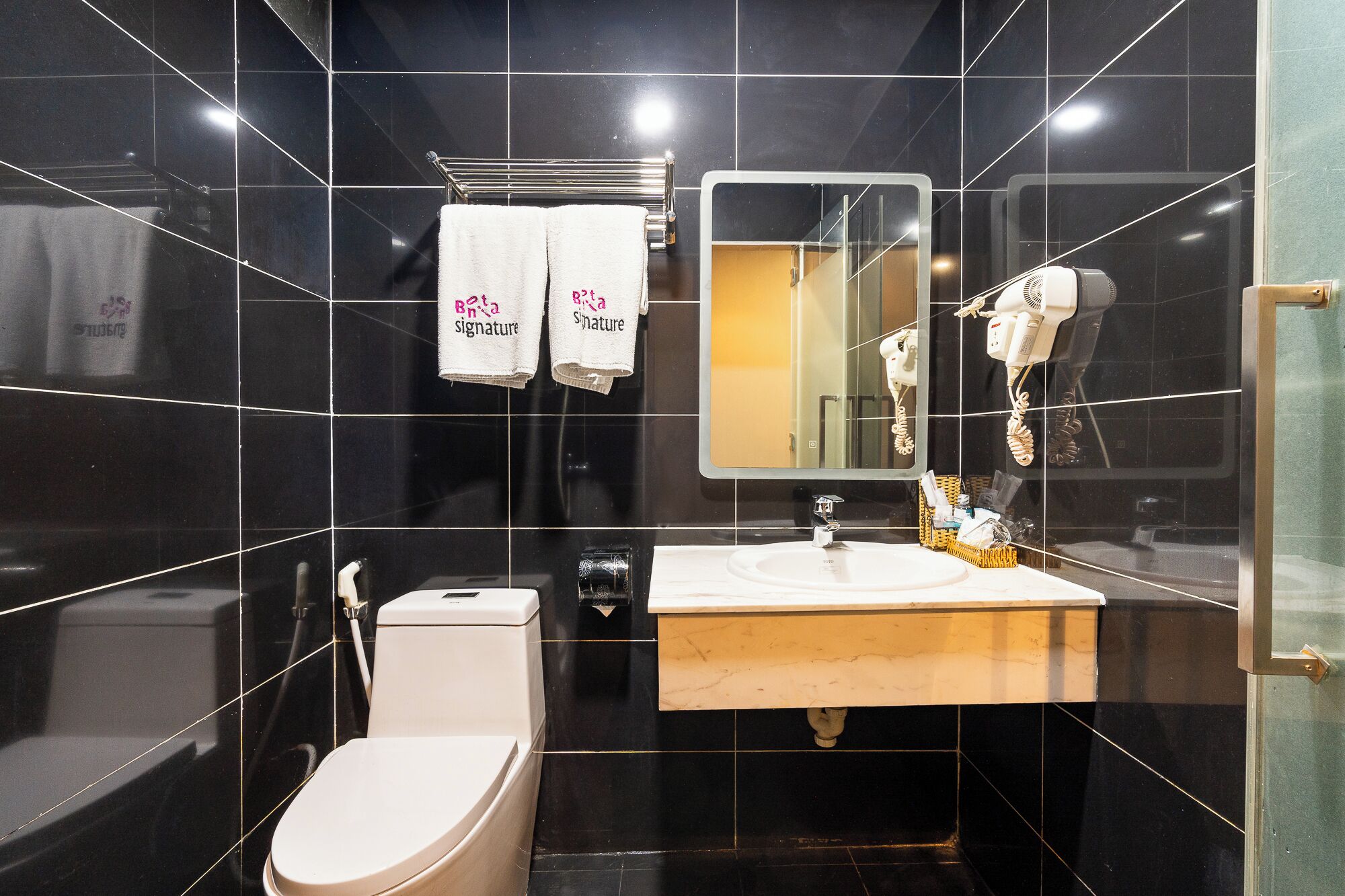 A modern bathroom with black tiled walls, a white toilet, a sink with a mirror above it, and a hairdryer on the wall.