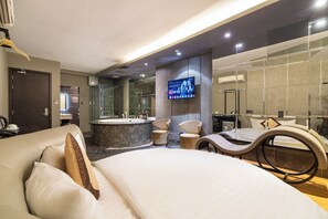 Una habitación de hotel moderna con una cama grande, un pequeño rincón de descanso con sillas, una bañera, un televisor montado en la pared y un espejo amplio.