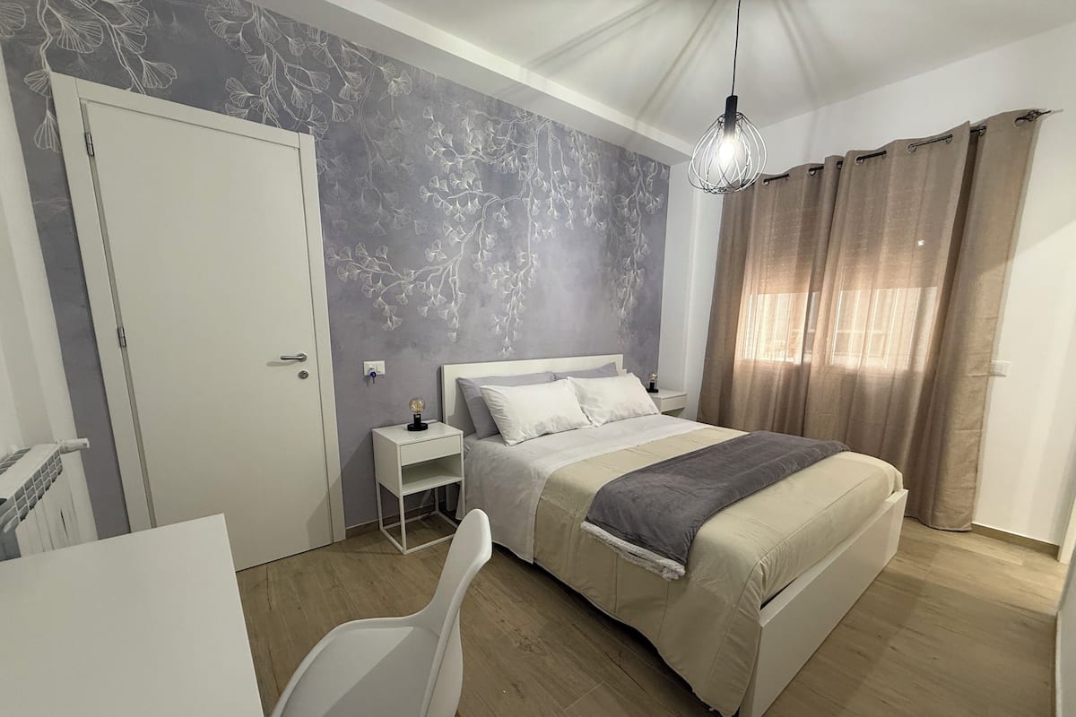 Une chambre à coucher comprenant un lit, une table de chevet, une chaise et un mur orné d’un motif de feuilles.