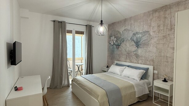 A bedroom with a bed, a desk, a TV, a balcony with curtains, and a wall with a floral mural. - Bart165 Luxury Rooms (Palermo)