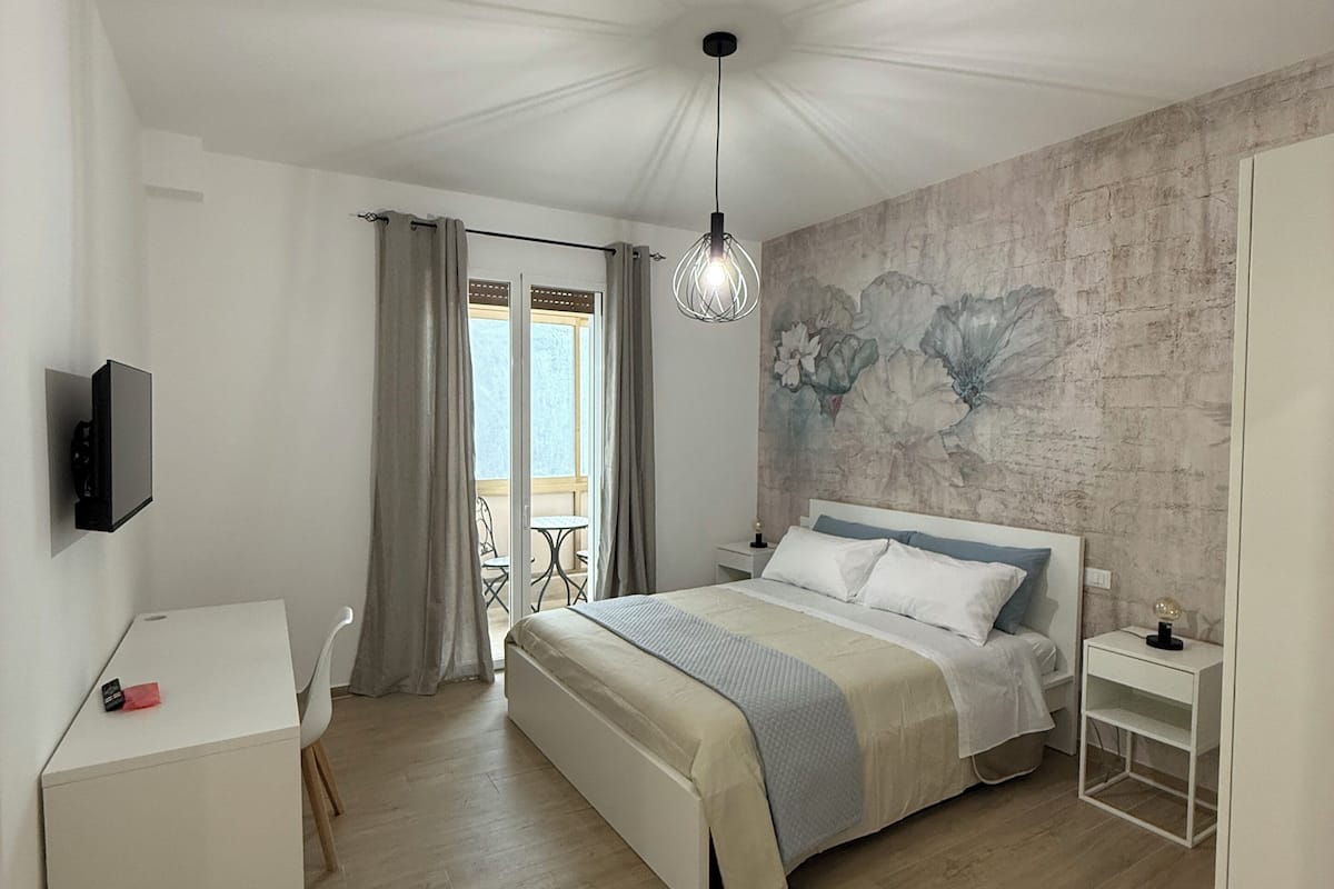 Une chambre à coucher comprenant un lit, un bureau, une télévision, un balcon avec des rideaux et un mur orné d’une fresque florale.