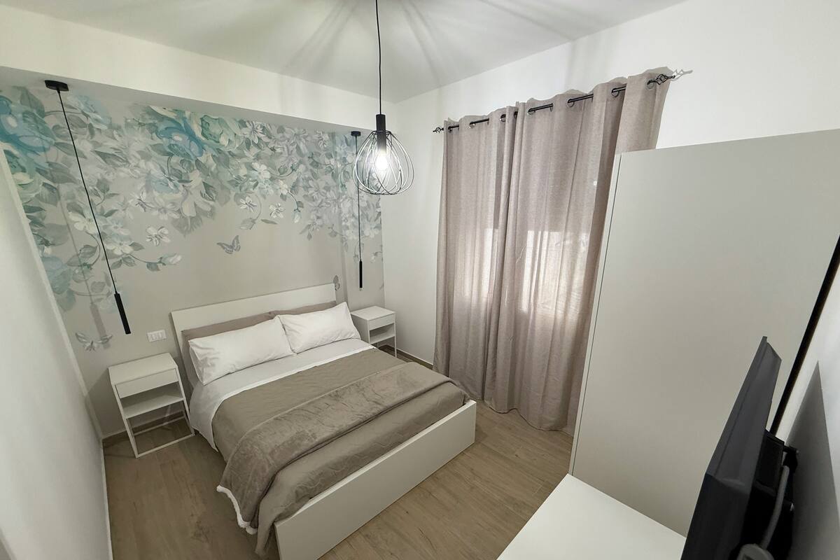 Une chambre à coucher comprenant un lit, des tables de chevet, un luminaire fixé au mur, un téléviseur et une fenêtre avec des rideaux.