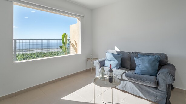 Ein modernes Wohnzimmer mit einem grauen Sofa, einem kleinen runden Tisch und einem großen Fenster mit Blick auf einen Balkon und eine entfernte Landschaft.