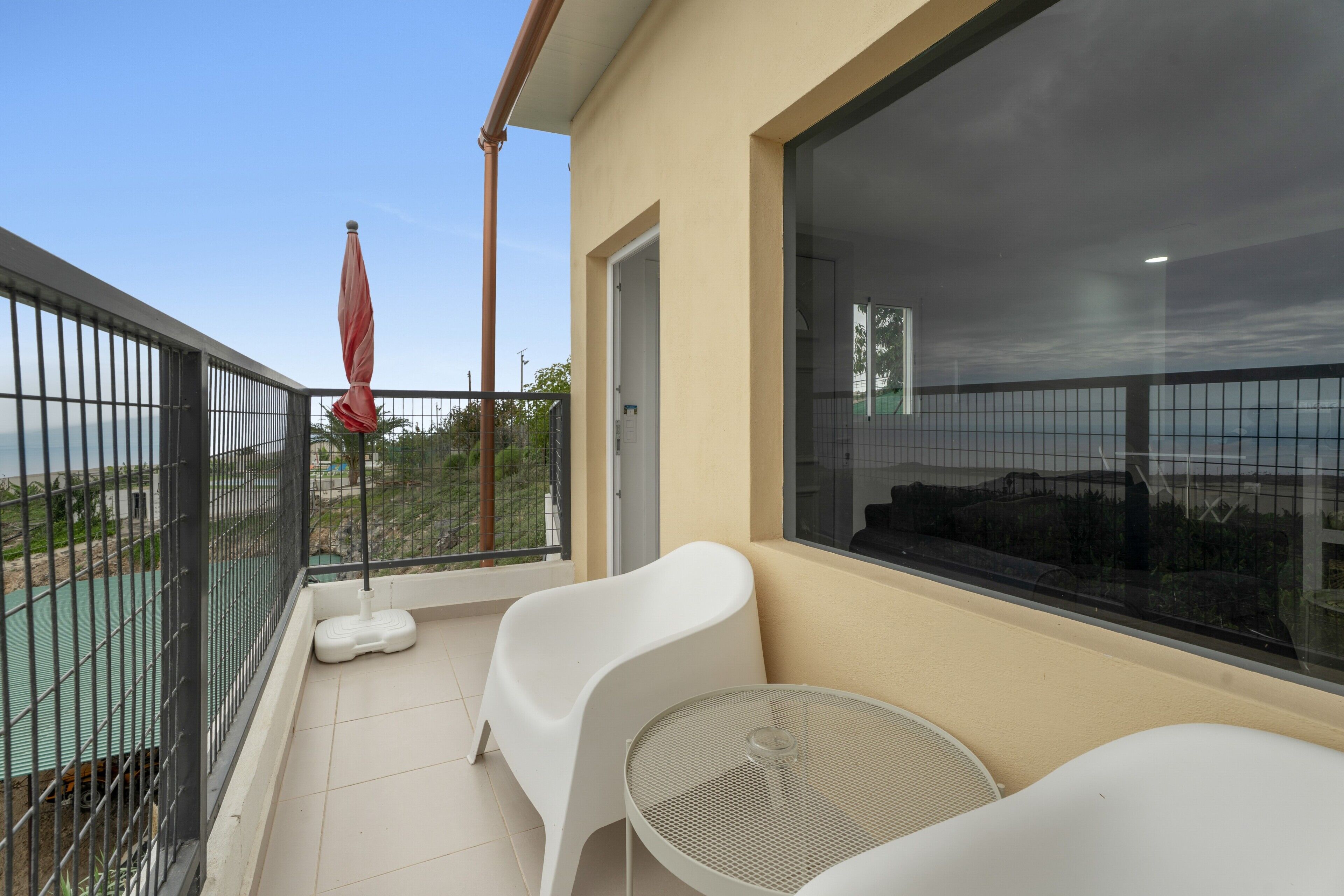 Ein Balkon mit einem Metalltisch, zwei weißen Stühlen, einem roten Sonnenschirm und Blick aufs Meer.
