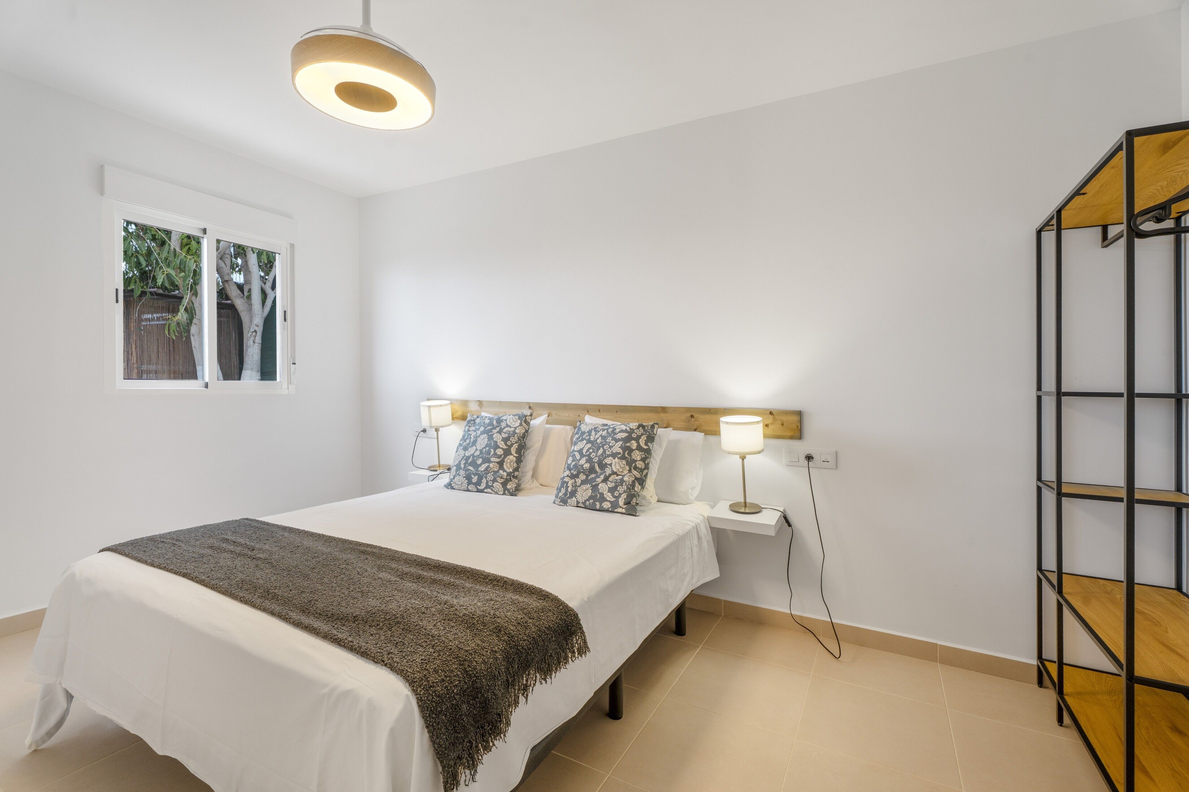 A bedroom with a bed, bedside lamps, a window, and a wooden shelf unit.