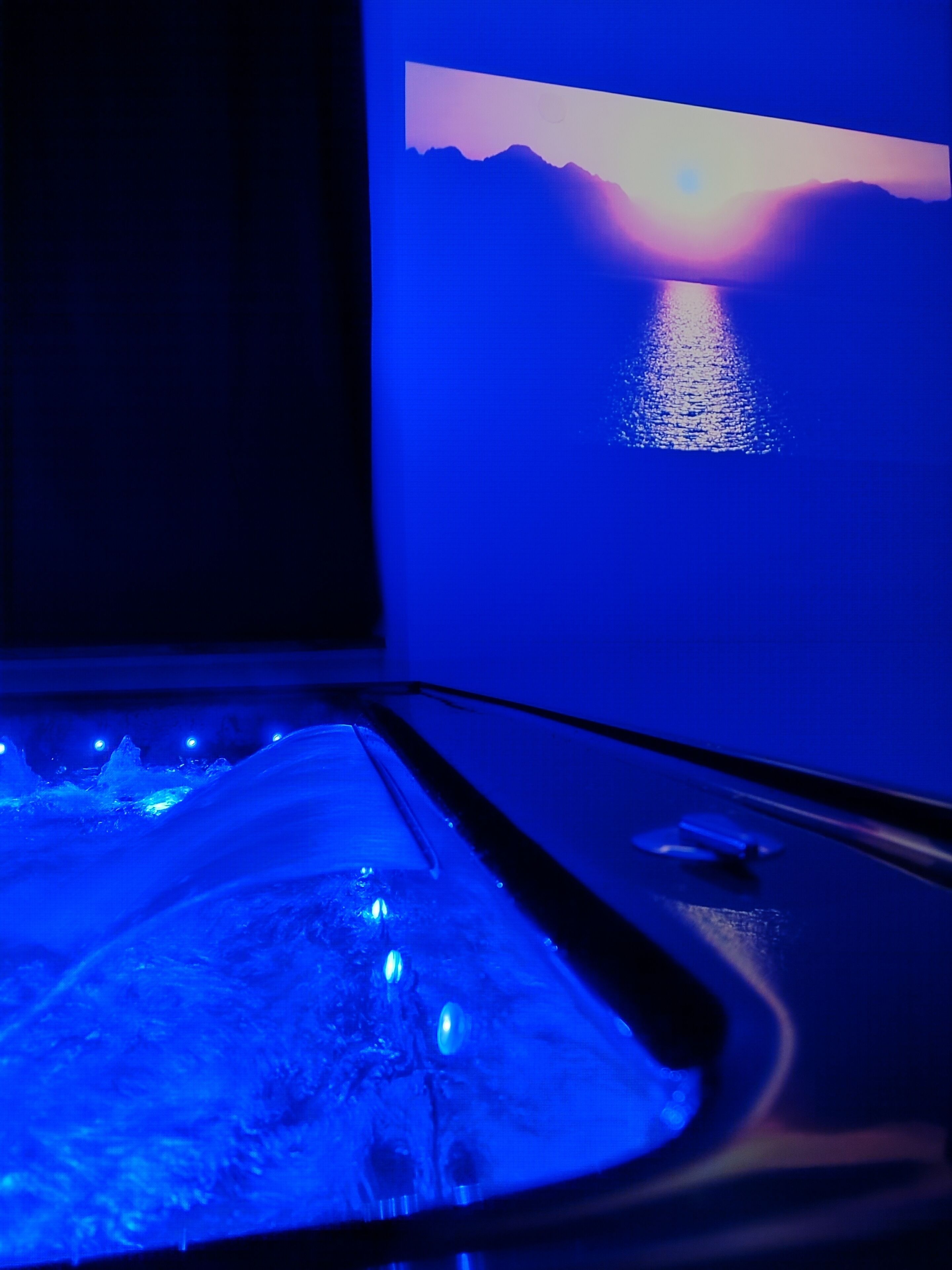 A room with a television displaying a sunset over water, a bed with a blue light under it, and a window with a view of mountains.