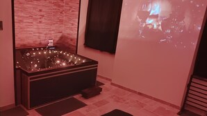 Um spa moderno com uma banheira grande, uma tela de projeção e um arranjo de pétalas de rosas em formato de coração no chão.