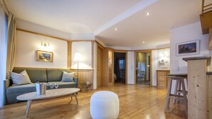 A modern living room with a sofa, a coffee table, and a round ottoman. There is a wooden floor and a kitchen area in the background. - 1010A Marienstern Apartments (Santa Cristina Valgardena)