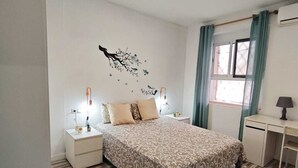 A bedroom with a bed, nightstands, a desk, a window with curtains, and a wall decoration. - Malvarrosa Beach | 3hab| 5hues| Pet friendly (València)