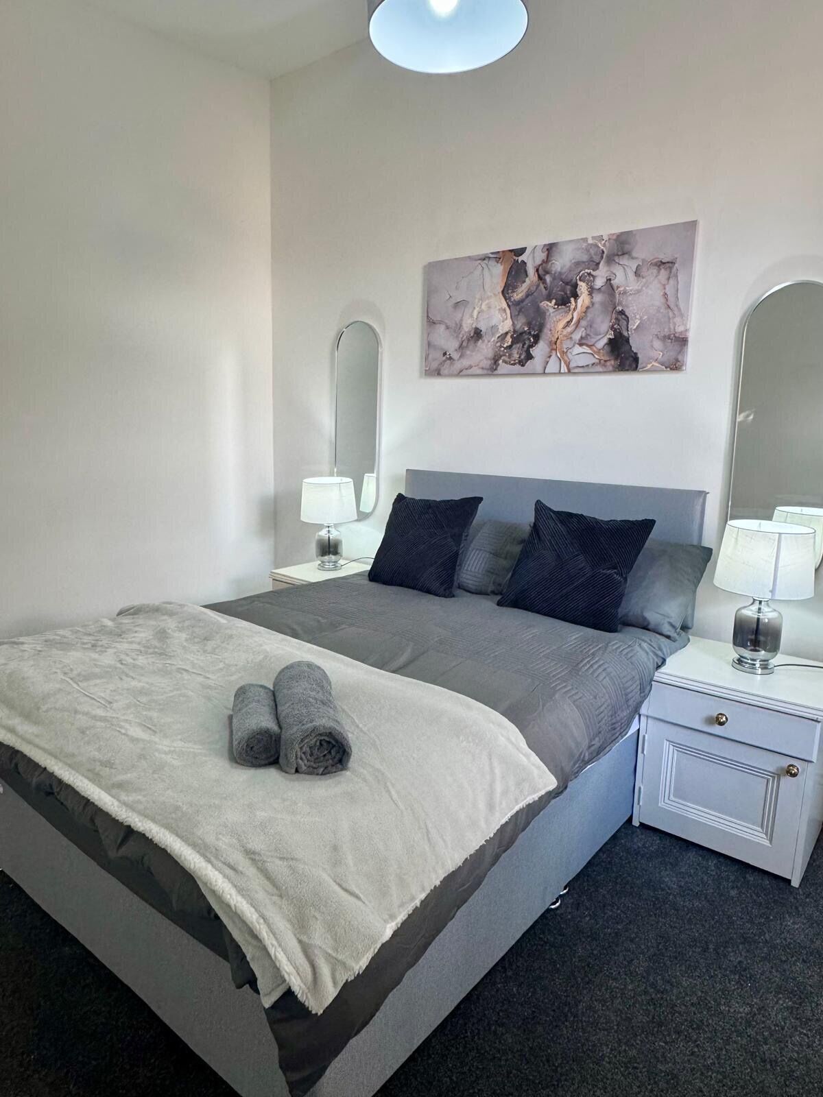 A neatly arranged bedroom with a bed, two bedside tables, a wall-mounted mirror, and a painting.
