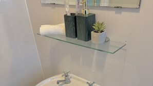 A bathroom with a white sink, two faucets, and a glass shelf above it holding toiletries. - Sleeps4- Sofabed- 5mintocitycentre- Glasgowgreen (Glasgow)