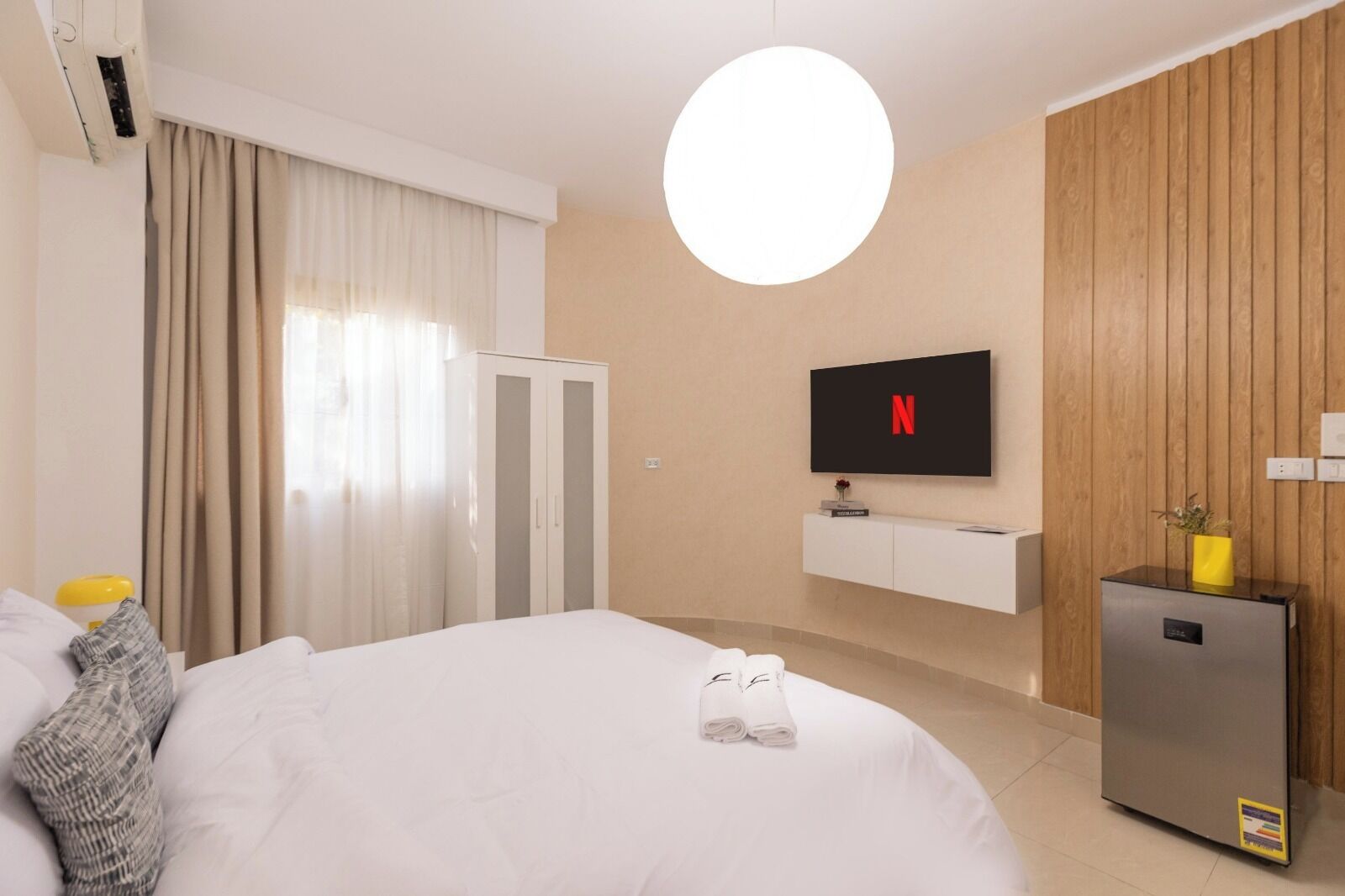 Quarto de hotel com cama, televisão na parede, minibar e painel de parede de madeira.