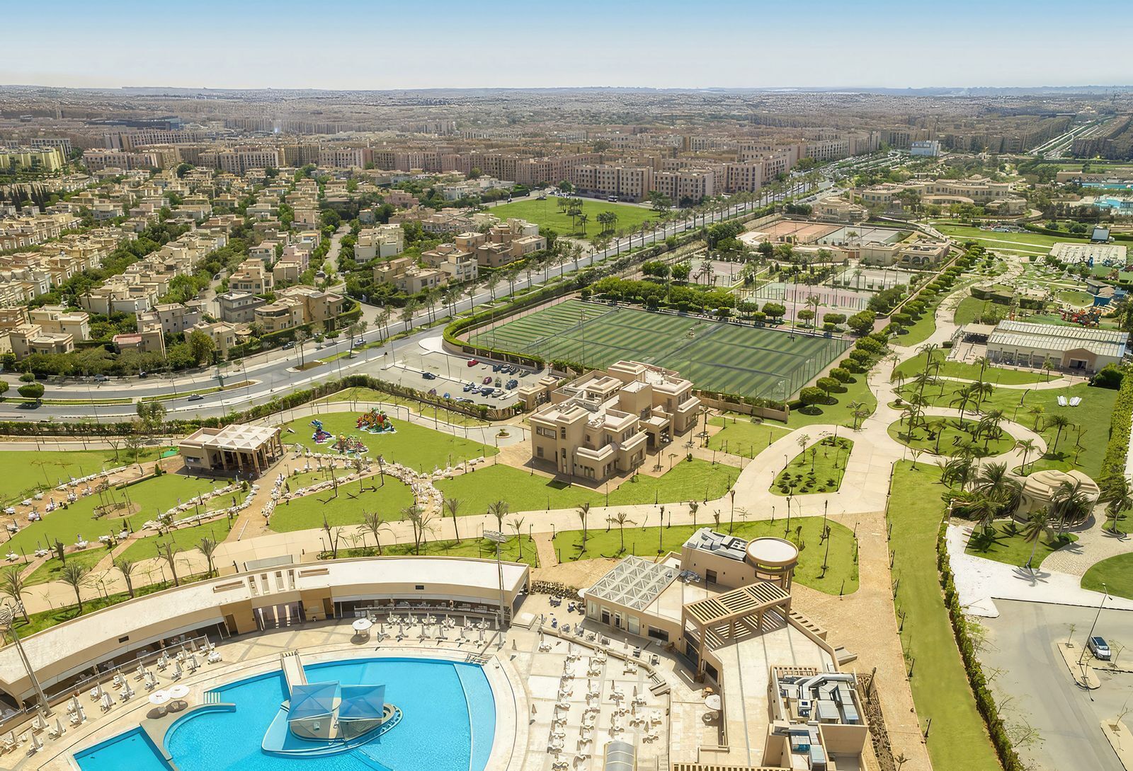 Vue aérienne d’un quartier résidentiel avec une grande piscine, un terrain de sport et un parc planté de palmiers.