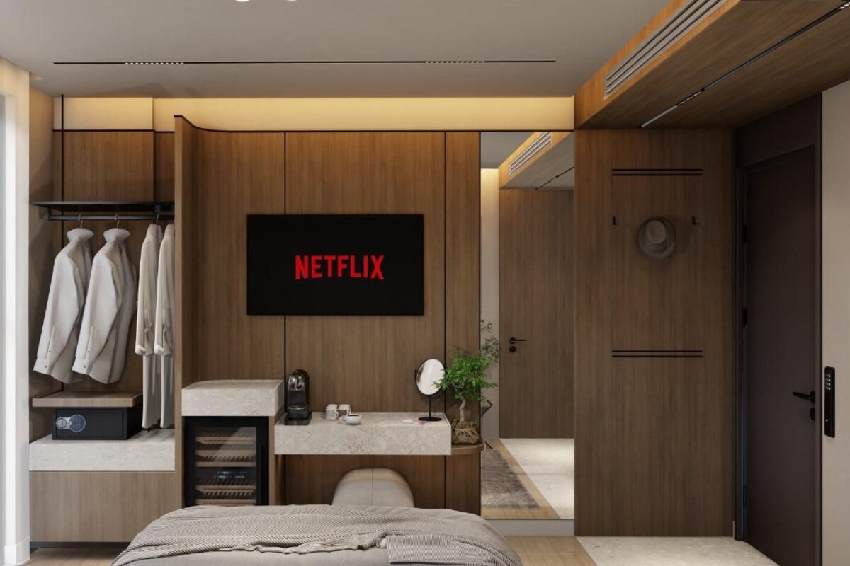 A modern bedroom with a large bed, a vanity with a mirror, a shelf with books, a wardrobe, and a television displaying the Netflix logo.
