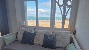 A white sofa with patterned cushions, a window with a view of the beach and ocean, and a lamp on the windowsill.