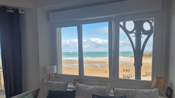 A room with a large window overlooking a beach, a sofa with patterned cushions, and a lamp on a side table.
