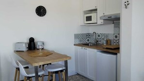 A compact kitchen with a wooden table, white chairs, a microwave, a sink, and a clock on the wall.