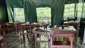 Un'area ristoro con tende, tavoli e sedie in legno, pavimento piastrellato e vista sul verde circostante.