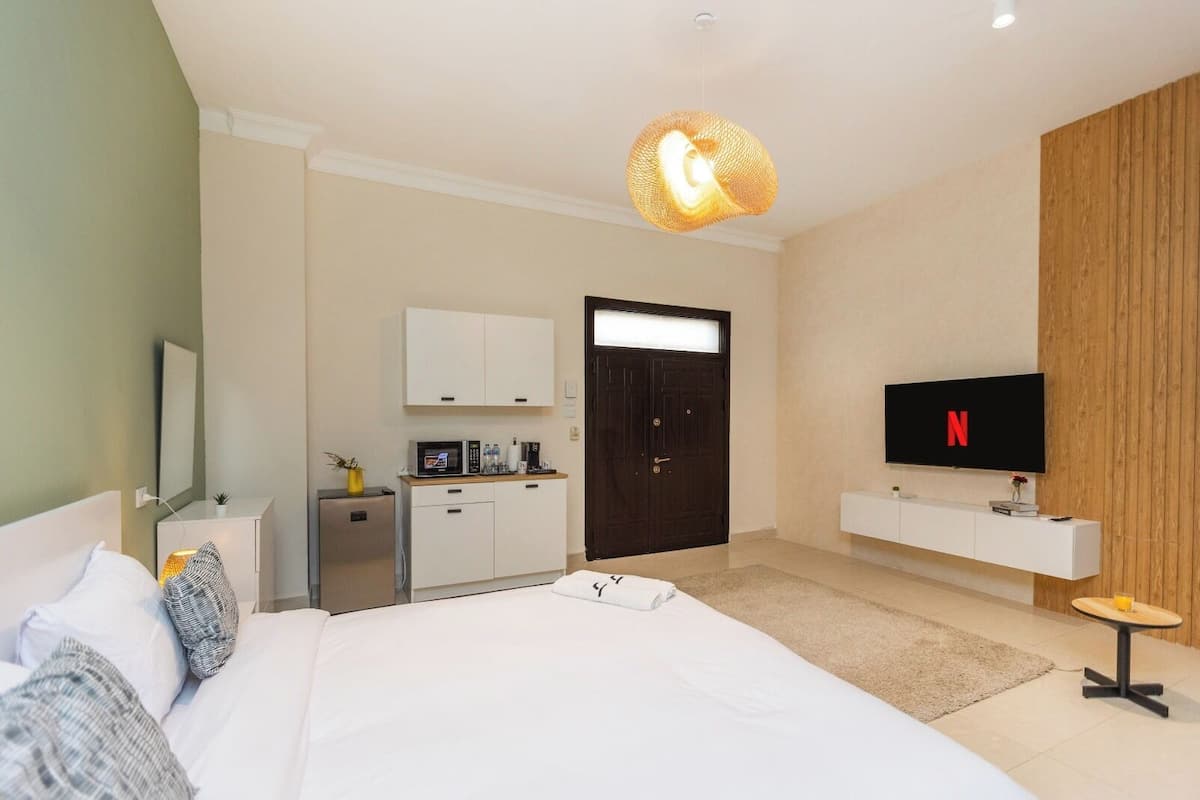 A modern hotel room with a large bed, a TV mounted on the wall, a minibar, and a wooden panel wall.