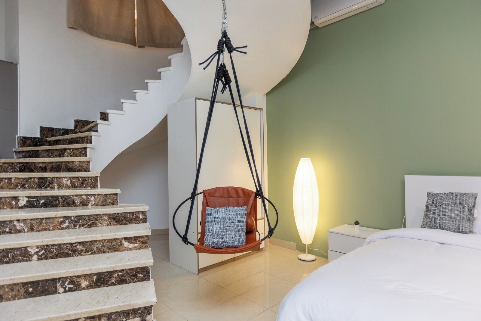 Ein modernes Hotelzimmer mit einer Marmortreppe, einem Hängekorbstuhl, einer Nachttischlampe und einem Bett mit weißem Kopfteil.
