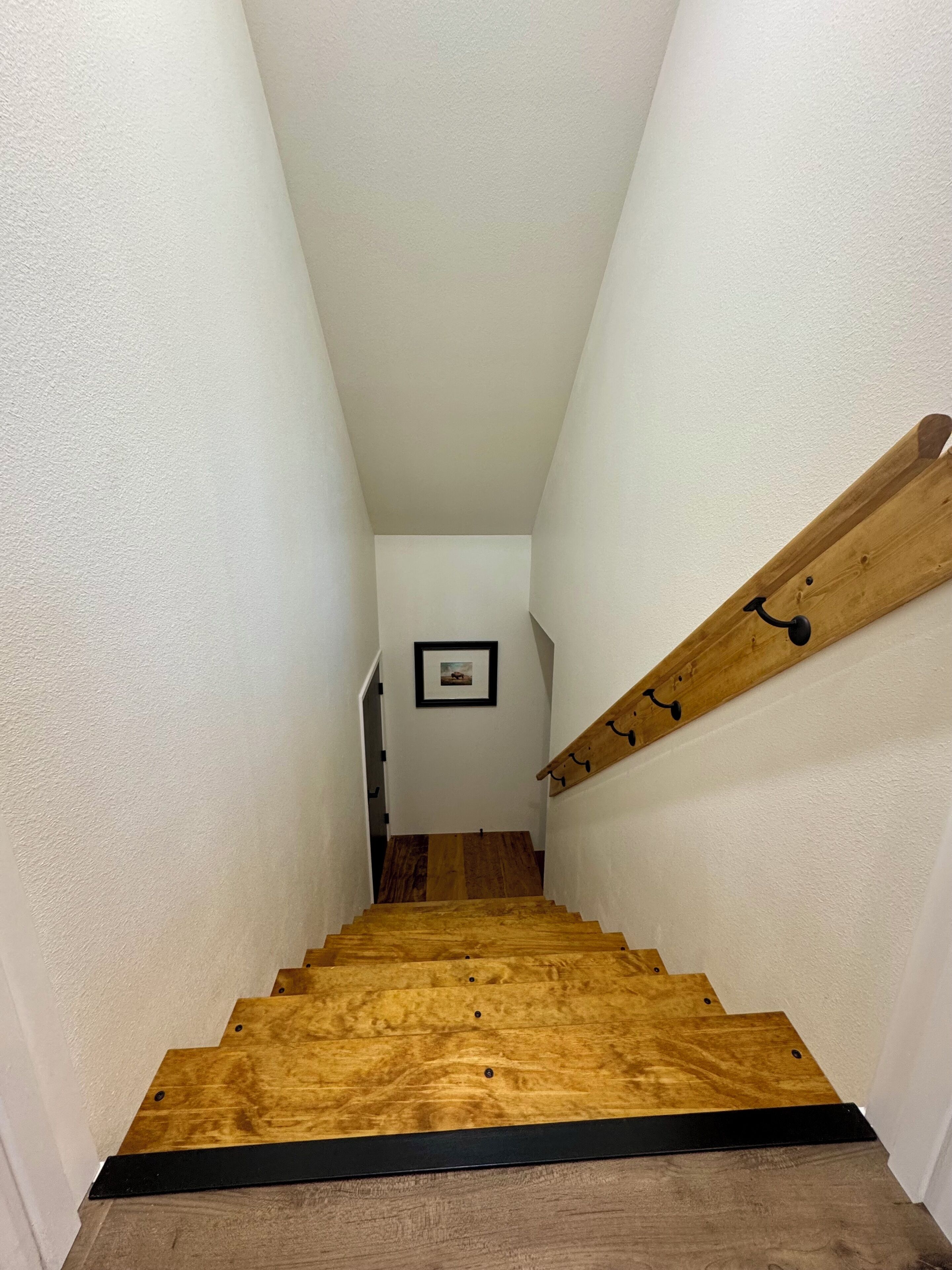 A narrow staircase with wooden steps and a black handrail, leading to a landing with a framed picture.