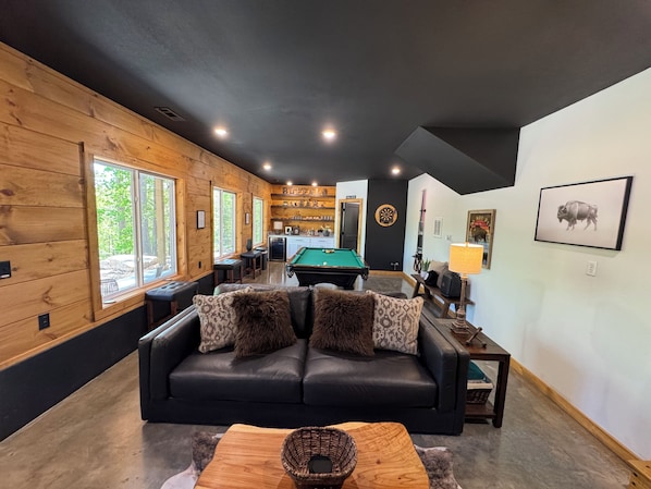 A cozy living room with a black leather sofa, a pool table, and a dining area with wooden walls and a kitchen in the background.