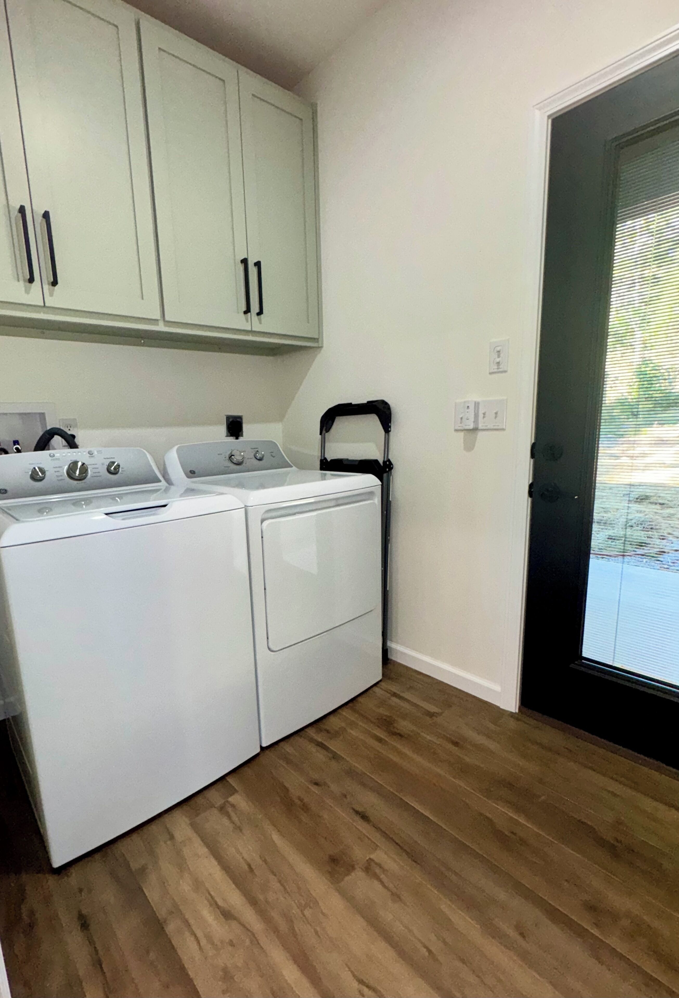 A laundry area with two white washers and dryers, wooden flooring, and light-colored cabinets.