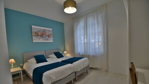 Un lit double avec une tête de lit, une table de chevet avec une lampe, une fenêtre avec des rideaux légers et un tableau accroché au mur.